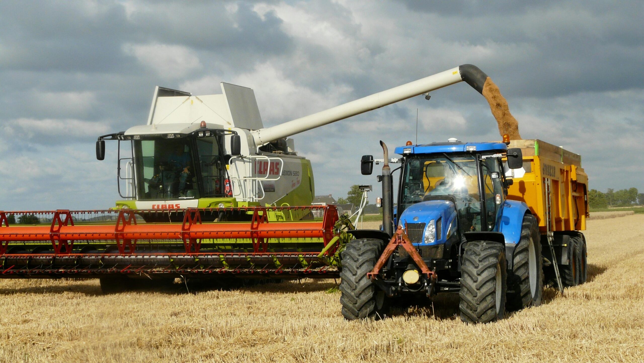 Home A modern combine harvester and tractor working together to harvest wheat in a sunny outdoor field.