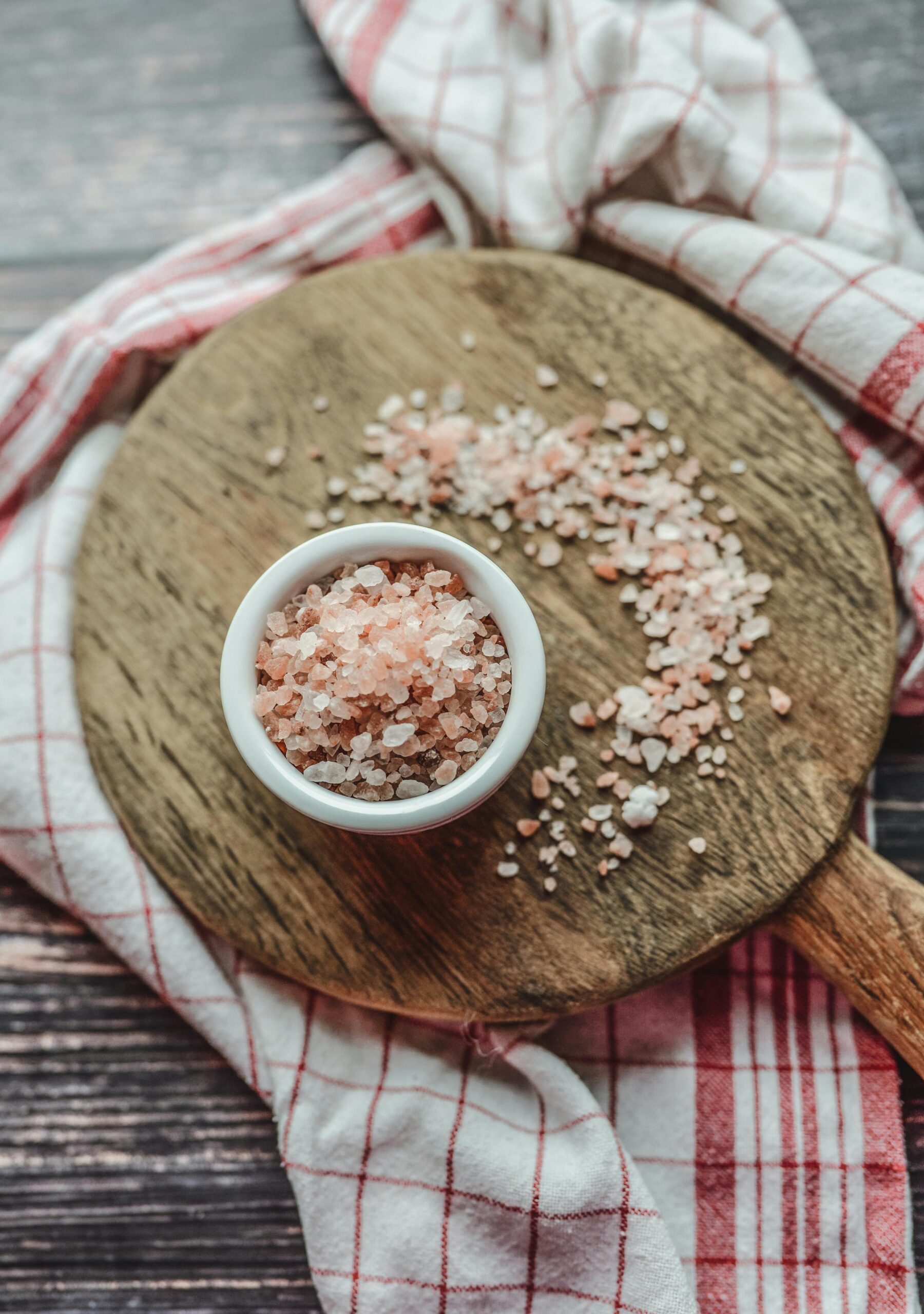 Home A bowl of pink Himalayan salt on a wooden board surrounded by a checkered cloth, viewed from above.