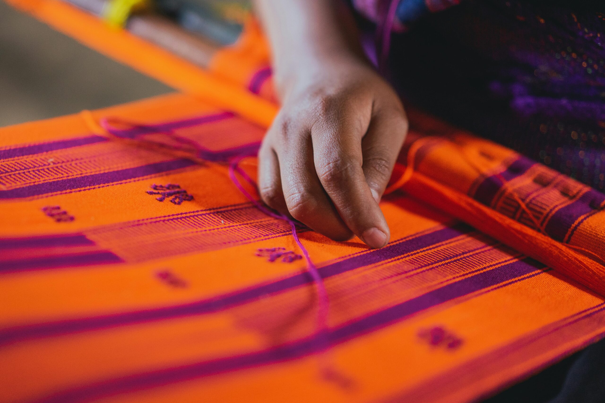 Home Close-up of a hand weaving a vibrant orange and purple textile, showcasing traditional craftsmanship.